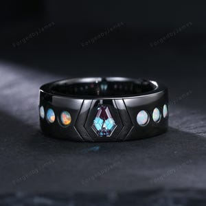 May include: A black ring with a geometric design. The ring features a central, multi-faceted, blue-green gemstone and several smaller, round, iridescent stones. The ring is set against a dark background.