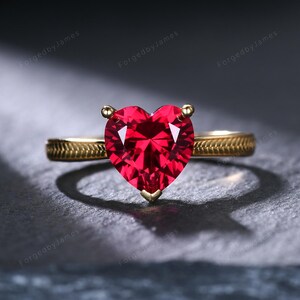 May include: A gold ring featuring a prominent heart-shaped red gemstone set in the center. The band of the ring displays a textured, etched pattern along its surface. The design includes three small prongs securing the vibrant red heart stone.