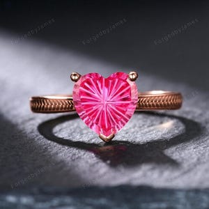 May include: A rose gold ring featuring a heart-shaped, faceted pink gemstone as the centerpiece. The metal band displays a textured, etched pattern along its sides, leading up to the prong setting that secures the vibrant, starburst-cut stone.