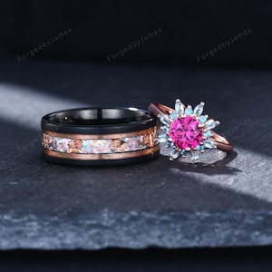 May include: A black band ring with a central inlay of iridescent flakes and rose gold accents, paired with a rose gold ring featuring a large round fuchsia center stone surrounded by a halo of smaller light blue marquise-cut stones.