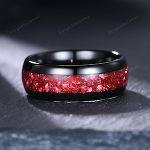 Puede incluir: Un anillo negro con una banda de piedra triturada roja vibrante. El anillo tiene un acabado pulido y brillante. El texto "ForgedbyJames" es visible en el fondo.