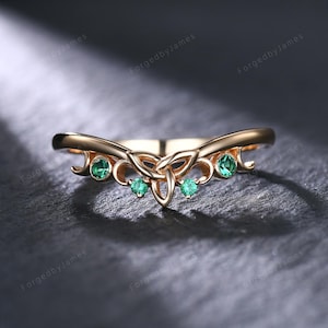 May include: A gold-colored ring with a Celtic knot design and emerald-green gemstones. The ring has a curved shape with three gemstones in the center and one on each side. The ring is set against a dark background.