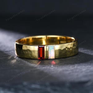 May include: A gold-toned ring with a hammered texture, featuring two rectangular baguette-cut stones set side-by-side: one deep red garnet and one iridescent white opal. The band has a polished finish and a consistent width throughout its design.