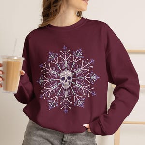 May include: A maroon long-sleeve crewneck sweatshirt featuring a central graphic of a human skull surrounded by a symmetrical, snowflake-like pattern composed of white and light purple bones and decorative fleur-de-lis elements.