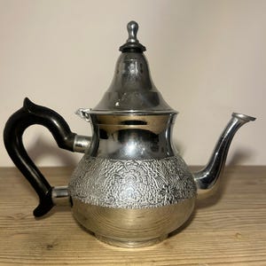 Puede incluir: Tetera plateada con asa y boquilla negras. La tetera tiene un diseño floral ornamentado alrededor del centro y una tapa cónica con un pequeño pomo en la parte superior. Está sobre una superficie de madera.