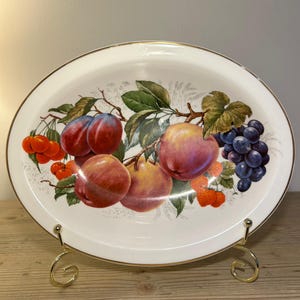Puede incluir: Plato ovalado de cerámica con diseño de frutas. El plato presenta un fondo blanco con un borde dorado y una colorida ilustración de ciruelas, manzanas, cerezas, uvas y fresas. El plato se exhibe sobre un soporte de metal dorado.