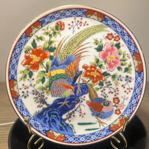 Puede incluir: Un plato decorativo de porcelana con un diseño colorido de pájaros y flores. El plato tiene una base blanca con un borde azul y rojo. El pájaro tiene plumas azules, verdes y naranjas, y las flores son rojas y rosas.