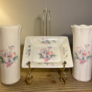 Può includere: Un set decorativo color crema con due vasi e un piatto quadrato, tutti adornati con un motivo floreale rosa, blu e verde. Il piatto è esposto su un supporto in metallo dorato. I vasi hanno un bordo smerlato.