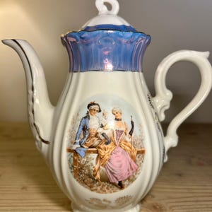 Könnte beinhalten: Weiße Porzellan-Teekanne mit blau-weißem Deckel und Griff. Die Teekanne zeigt eine gemalte Szene von zwei Figuren in historischer Kleidung. Goldene Verzierungen akzentuieren Ausguss und Griff und verleihen dem Design einen Hauch von Eleganz.