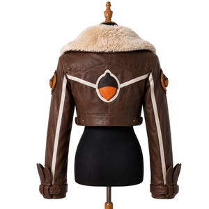 May include: A brown leather jacket featuring a large cream-colored faux fur collar, long sleeves with buckled cuffs, and a prominent acorn-shaped emblem on the back. The design includes white vertical stripes and additional circular accents.