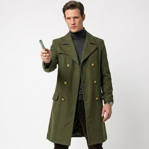 May include: Olive green double-breasted coat with gold buttons. The coat is worn over a gray turtleneck and brown corduroy pants. A silver watch is visible on the wrist. The person is holding a green and silver device.
