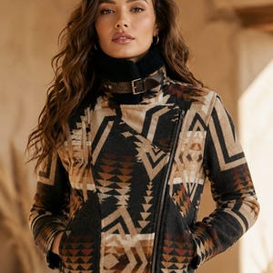 May include: A moto-style jacket featuring a geometric pattern in shades of black, tan, and rust. The jacket has a black faux-shearling collar with a brown leather strap and buckle closure, a diagonal front zipper, and two side pockets, worn with blue jeans.
