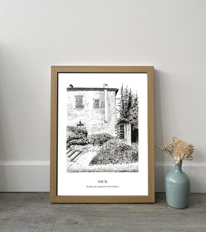 May include: Framed black and white print of a building in Nice, France, with the text "NICE Jardin du monast&egrave;re de Cimiez". The print is in a light brown wooden frame and is displayed on a gray wooden surface.