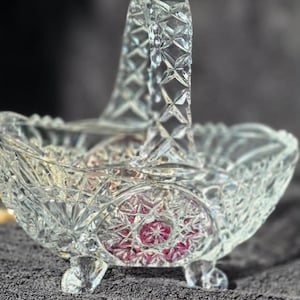 Może przedstawiać: Kosz z przezroczystego szkła o skomplikowanym wzorze szlifu diamentowego z wysokim, łukowatym uchwytem. Podstawa kosza wsparta jest na trzech małych nóżkach, a boki zdobi wyraźny okrągły medalion z ciemnoróżowym wzorem w kształcie gwiazdy.