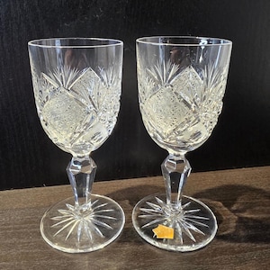 May include: A pair of clear crystal wine glasses, each with detailed cut glass patterns. The design includes diamond shapes and starbursts. The faceted stems and bases are visible. The glasses are displayed on a dark surface.