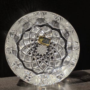 May include: A clear, round lead crystal ashtray with a starburst design in the center. The ashtray features a geometric pattern and a textured rim. A small label is affixed to the center. The ashtray is hand-cut.