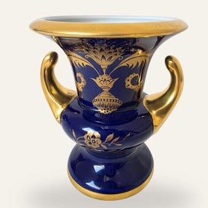 Może przedstawiać: Głęboko niebieski wazon ceramiczny z szerokim, złoconym otworem i dwoma wydatnymi uchwytami w kolorze złota. Zewnętrzna strona ozdobiona jest misternymi złotymi wzorami kwiatowymi i roślinnymi, a podstawa wykończona jest jednolitym złotym pasem.