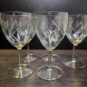 May include: A set of four clear glass goblets with stems and circular bases. Each glass features a delicate etched pattern of intersecting diagonal lines and small circular accents around the bowl, creating a light and airy visual texture.