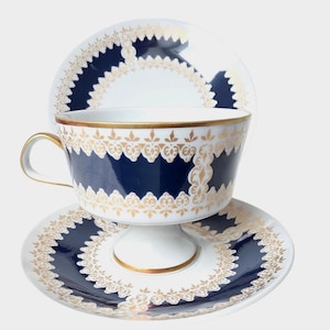 Może przedstawiać: Zestaw filiżanki ze spodkiem z białej porcelany, ozdobiony głębokim granatowym pasem ze skomplikowanym złotym wzorem filigranowym. Filiżanka posiada podstawę w formie cokołu i złocone ucho, a spodek wykazuje pasujący, falisty wzór na obrzeżach.