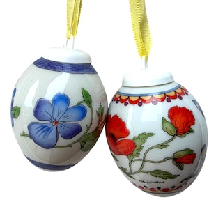 May include: Two white ceramic egg-shaped ornaments hanging from yellow ribbons. One egg features a large blue flower with green leaves and blue bands at the top and bottom. The other egg displays red poppy flowers with green stems and a decorative scalloped border.