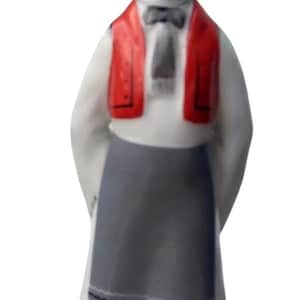 Può includere: Una statuetta in porcellana raffigurante una persona con un cilindro nero con fascia floreale, camicia bianca, gilet rosso, grembiule grigio con bordo floreale e scarpe nere. La figura poggia su una base bianca con le mani dietro la schiena.