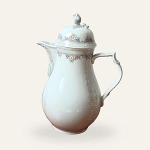 May include: A white porcelain coffee pot with a lid and handle. The pot is decorated with a floral pattern in pastel colours. The lid is topped with a small rose bud. The handle has a decorative design.