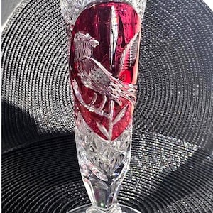 May include: A clear and ruby red cut glass vase featuring a scalloped rim, a diamond-patterned body, and a central panel with an etched bird design. The base is circular with a starburst pattern and a small circular sticker on the surface.