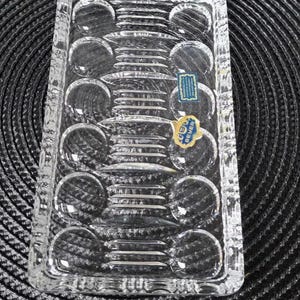May include: A rectangular clear crystal tray featuring a series of circular indentations along the sides and horizontal ridges in the centre. Two blue and gold labels are attached to the surface, one reading Beyer, and the other with illegible text.