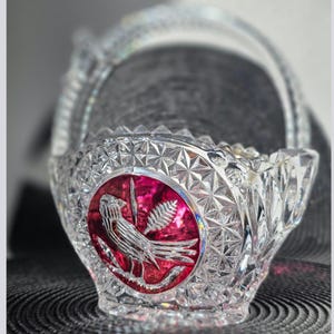 Puede incluir: Una cesta de vidrio prensado transparente con un patrón de diamantes facetados y un medallón circular prominente en rojo oscuro. El medallón muestra un diseño en relieve de un pájaro posado en una rama con una hoja sobre el cuerpo de vidrio.