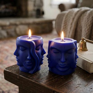 May include: Two blue candles with multiple faces carved into them, each with a lit wick. The candles are placed on a dark wooden table. The background includes a fireplace, a couch, and a bell.