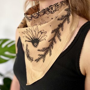 May include: A tan bandana with a black floral print featuring a daisy and pine branches. The bandana is tied around a person's neck.