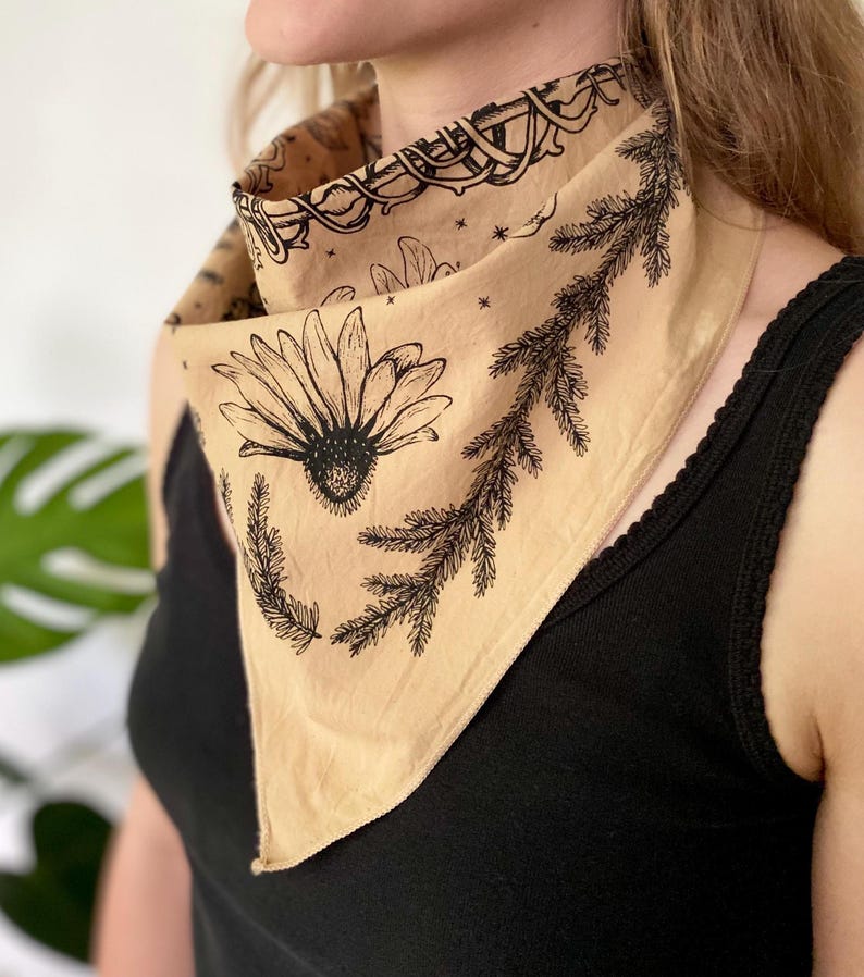 May include: A tan bandana with a black floral print featuring a daisy and pine branches. The bandana is tied around a person's neck.