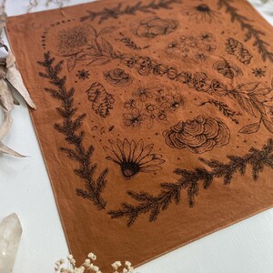 May include: A brown bandana with a black floral design. The design includes flowers, leaves, and other botanical elements. The bandana has a fringed edge.