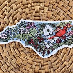 May include: A sticker in the shape of North Carolina, featuring a red cardinal bird, flowers, and foliage. The design includes white, pink, and red flowers, with a light blue background. The sticker is on a woven background.