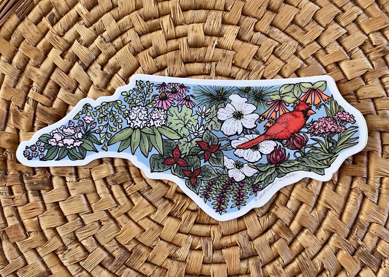 May include: A sticker in the shape of North Carolina, featuring a red cardinal bird, flowers, and foliage. The design includes white, pink, and red flowers, with a light blue background. The sticker is on a woven background.