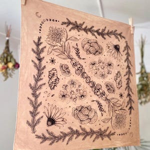 May include: A beige cotton bandana with a black ink illustration of various plants, flowers, and mushrooms. The design is centered in a square frame with a crescent moon and stars in the corners.