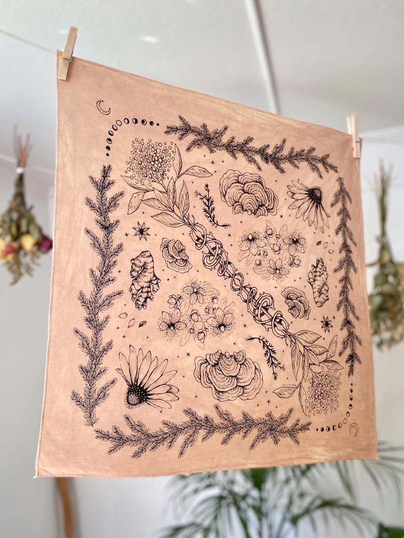 May include: A beige cotton bandana with a black ink illustration of various plants, flowers, and mushrooms. The design is centered in a square frame with a crescent moon and stars in the corners.