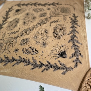 May include: A tan bandana with a black floral and botanical print. The print includes flowers, leaves, and stars. The bandana is made of a soft, lightweight fabric.