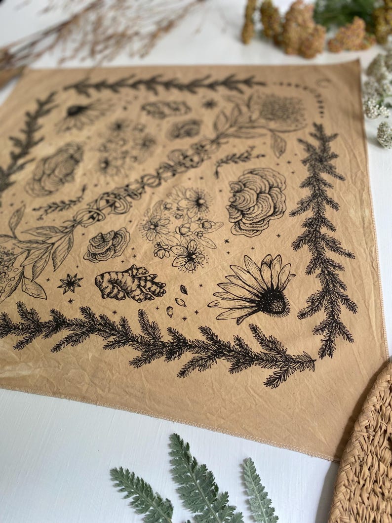 May include: A tan bandana with a black floral and botanical print. The print includes flowers, leaves, and stars. The bandana is made of a soft, lightweight fabric.