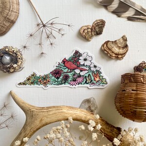 May include: A sticker depicting a red cardinal bird perched on a branch surrounded by colourful flowers, forming the shape of the state of Virginia. The sticker is on a white background.