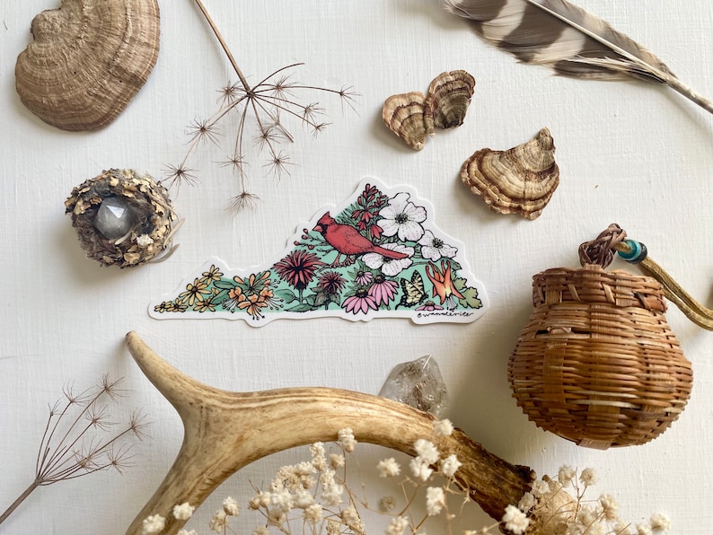 May include: A sticker depicting a red cardinal bird perched on a branch surrounded by colourful flowers, forming the shape of the state of Virginia. The sticker is on a white background.