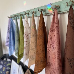May include: A set of eight tea towels hanging on a rustic wooden rack. The towels are made of natural fabric and feature a variety of botanical illustrations in brown, pink, and green hues.
