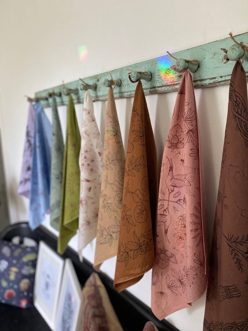 May include: A set of eight tea towels hanging on a rustic wooden rack. The towels are made of natural fabric and feature a variety of botanical illustrations in brown, pink, and green hues.