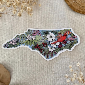 May include: A sticker in the shape of North Carolina, filled with colorful flowers and greenery. A bright red cardinal bird is perched on a branch. The background is light blue.