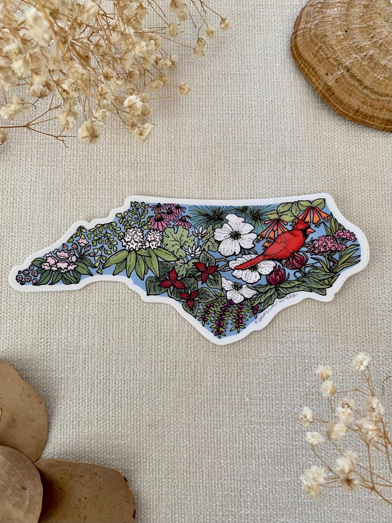 May include: A sticker in the shape of North Carolina, filled with colorful flowers and greenery. A bright red cardinal bird is perched on a branch. The background is light blue.