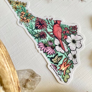May include: A sticker featuring a red cardinal bird perched on a branch with pink, white, and yellow flowers. The background is a light green watercolour wash. The sticker has the text "wanderlite" at the bottom.