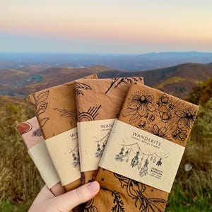 May include: Four brown fabric bandanas with botanical illustrations and the text "Wanderite Cosmic Design Studio" and "Herbalist Bandana" printed on them.