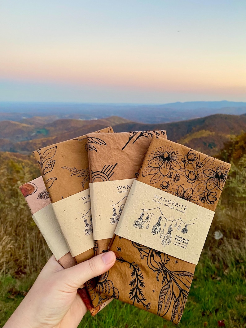 May include: Four brown fabric bandanas with botanical illustrations and the text "Wanderite Cosmic Design Studio" and "Herbalist Bandana" printed on them.