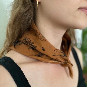 May include: A person wearing a brown bandana with a black floral design around their neck. The bandana has a white border. The person is wearing a black tank top.