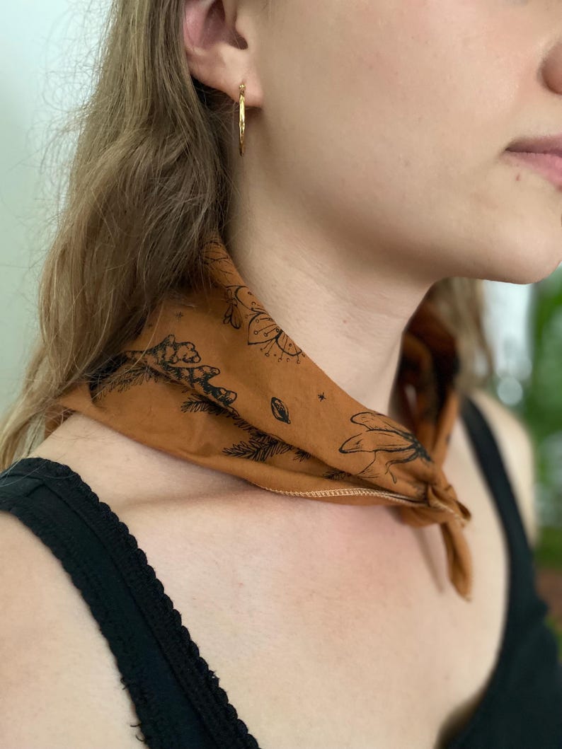 May include: A person wearing a brown bandana with a black floral design around their neck. The bandana has a white border. The person is wearing a black tank top.